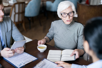 Serious female leader sitting with notebook and cup of tea and giving instructions to her...
