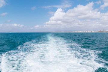 View from the ship on rough waves and the shore going away. A beautiful, sunny day at sea, in the distance, bright Mediterranean buildings. A bright, clear sky with white clouds.