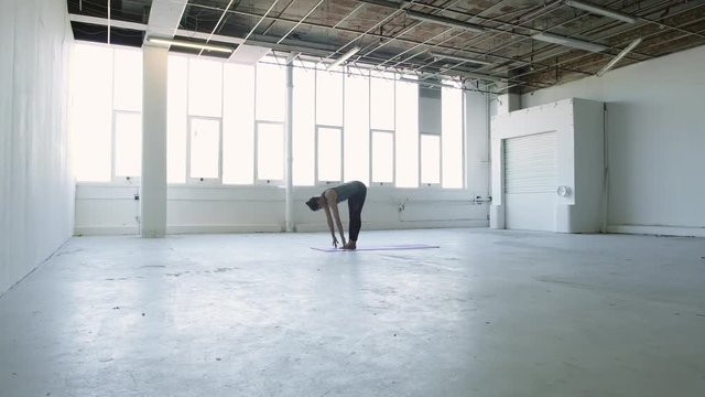Wide moving shot of a young brunette woman practicing yoga doing part of a sun salutation in a white warehouse studio.