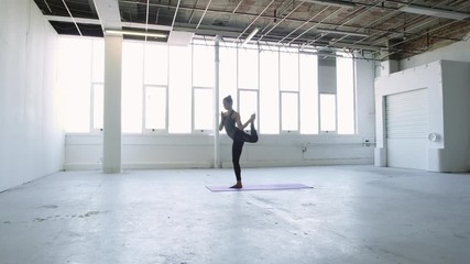 Wide orbiting shot of a flexible young brunette woman practicing yoga doing an advanced balance dancers pose in a white warehouse studio.