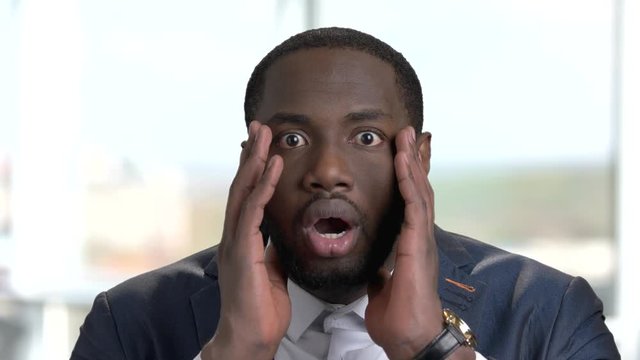 Portrait of shocked african american in suit. Amazed and surprised black business man against windows background.