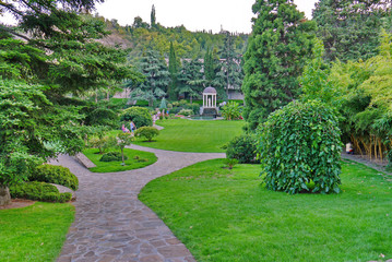 A beautiful view of the green lawns in the park with the paths running between them and people resting on the benches.
