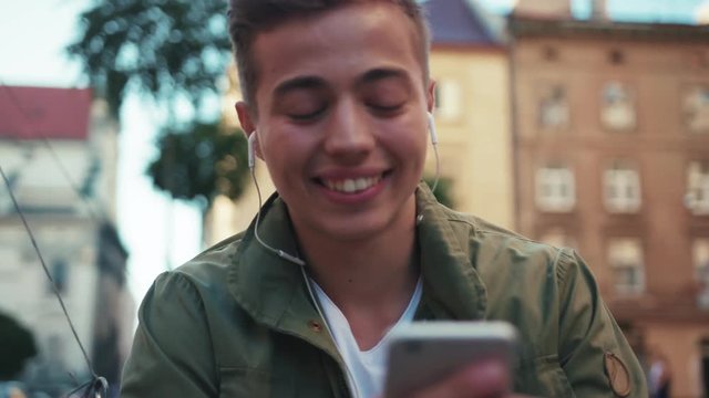face happy handsome young man use phone listen music in headphone look around sitting on steet smile cell sunlight mobile gadget technology hipster slow motion portrait