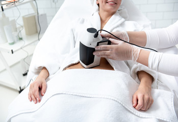 Obraz premium Outgoing lady having massage of stomach with special digital device while lying on couch. Cosmetologist holding it in arms