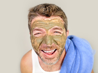 young attractive and happy man with bathroom towel  laughing of himself in the mirror with green cream on his face applying facial mask