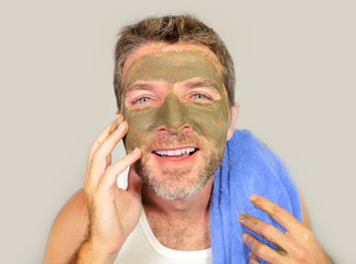 lifestyle isolated portrait of young attractive and happy man with bathroom towel  laughing of himself in the mirror with green cream on his face applying