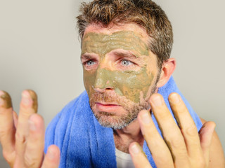 young messy funny man looking at himself horrified in bathroom mirror with green cream on his face applying beauty facial mask product