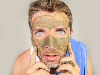 young messy funny man looking at himself horrified in bathroom mirror with green cream on his face applying beauty facial mask product