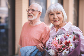 Waist up portrait of smiling senior lady strolling with male outside. She is happily holding bunch of flowers and looking at camera with joy © Yakobchuk Olena