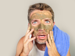 young messy funny man looking at himself horrified in bathroom mirror with green cream on his face applying beauty facial mask product