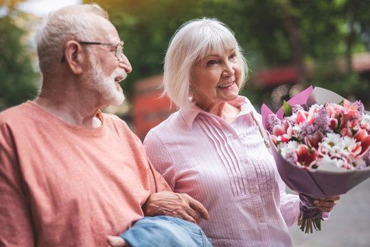 Side view of smiling man walking with his wife on street. Old lady is carrying bouquet of flowers with delight