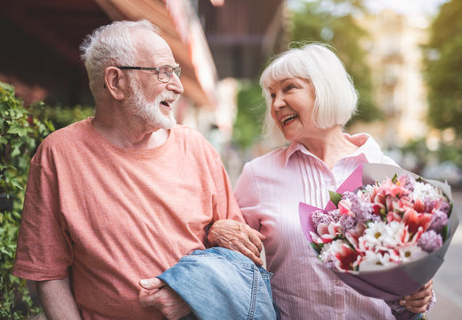 Side view of smiling senior male and female walking side by side outside after date. Mature woman is carrying bouquet of flowers and looking at man with sincere smile