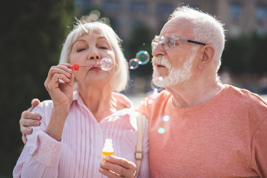 Back to childhood. Delighted senior man and woman standing on street and blowing bubbles with joy. Mature man is hugging wife while she is holding bottle with soap water