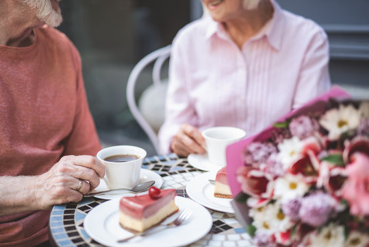 Delighted mature woman and man sitting at table outside and drinking tea with dessert. They are happily talking. Bunch of beautiful flowers is lying nearby