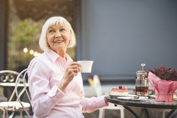 Fototapeta na wymiar Waist up portrait of smiling mature lady sitting at table with cup of tea in hands. She is looking sideways with joy while having piece of cake lying on table