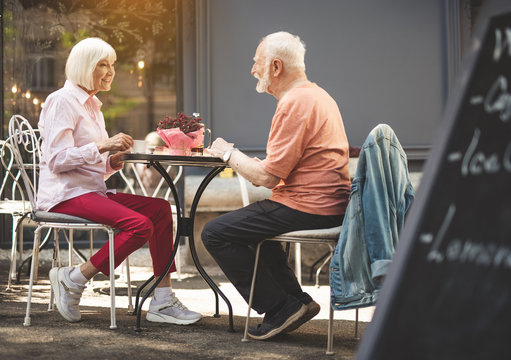 Profile full length of senior man and woman having date outdoors. They are sitting facing each other and drinking tea while taking with content