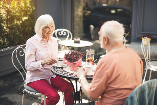 Full length of smiling old lady sitting at table and drinking tea with man. They are facing each other and looking with love and admire