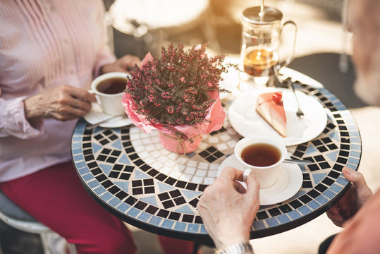 Senior couple sitting at table and drinking tea. They are holding cups and eating cake. Bunch of flowers is in the middle