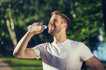 Side view of smiling male standing in park and holding bottle with liquid. He is happily refreshing during hot day