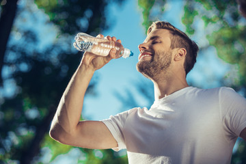 Low angle of happy sportsman drinking water outdoors. He is cheerful to satisfy thirst