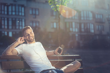 Waist up portrait of delighted man resting on bench with smartphone in hands. He is joyfully listening to music admiring beautiful morning outdoors. Copy space in right side
