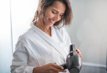 Portrait of optimistic woman looking at hairdryer while holding it in hands