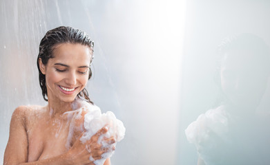 Portrait of beaming woman rubbing body with foam while standing under steam of water. Copy space