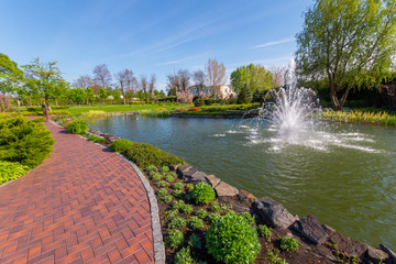 A path in the park passing next to a small pond with a fountain. With green lawns around and flowering trees.