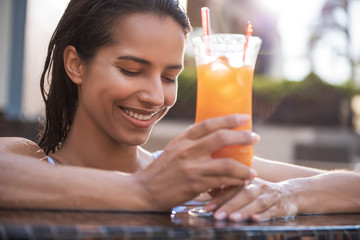 Satisfied woman with attractive smile tasting appetizing orange cocktail with ice. Her hair is wet