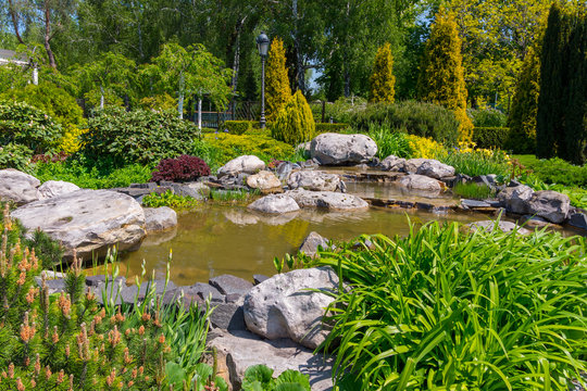decorative pond surrounded by boulders, flowers, green bushes, deciduous and coniferous trees