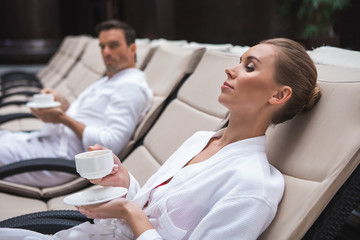 Side view focus on relaxed female lying on deckchair in bathrobe with cup of coffee in hands. Attractive man looking at her and sitting nearby is on background
