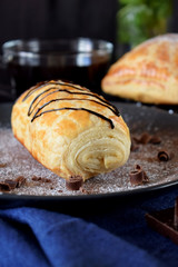 Puff pastry bun decorated with chocolate on a dark plate sprinkled with sugar powder