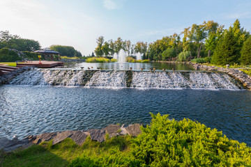 An artificial cascading waterfall flows from a lake in the park near a wooden gazebo with a terrace. Novi Petrivtsi, Ukraine