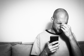 Young bald depressed man holding his head and cell phone with his hands feeling frustrated because he lost his job after his boss call him
