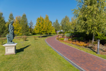 A statue of a woman with a thoughtfully lowered head against a blue sky standing on a green grass in a park next to a path going deep.