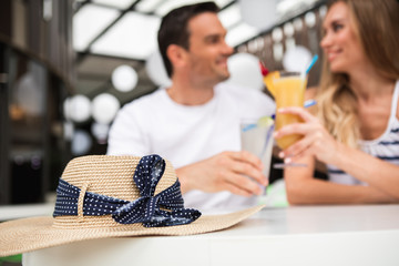 Focus on summer hat lying on table at cafe. Affectionate couple is having cocktail on background. They are clinking glasses and looking at each other with love