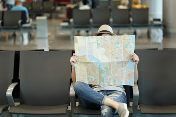 Young traveler tourist woman covering with paper map, search route, waiting in lobby hall at international airport. Passenger traveling abroad on weekends getaway. Air travel, flight journey concept.