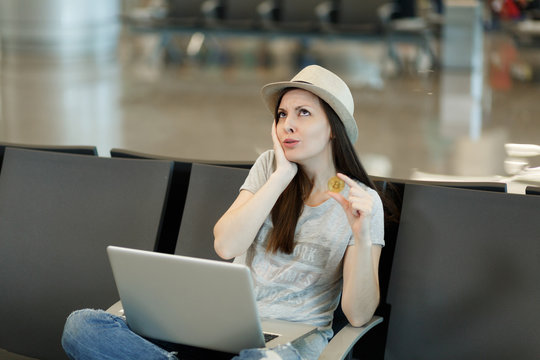 Young concerned traveler tourist woman working on laptop holding bitcoin, thinking and keeping hand near face, wait in lobby hall at airport. Passenger traveling abroad on weekend. Air flight concept.
