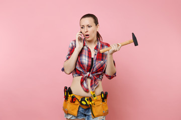 Angry handyman woman in plaid shirt, kit tools belt full of instruments talking on mobile phone, conducting unpleasant conversation isolated on pink background. Female in male work. Renovation concept