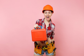 Strong young handyman woman in orange helmet, plaid shirt, denim shorts, kit tools belt full of instruments, toolbox isolated on pink background. Female in male work. Renovation and occupation concept