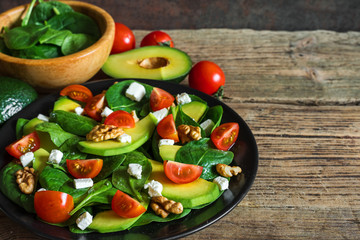 Fresh salad with avocado, spinach, tomatoes cherry, feta cheese and walnuts in a plate on rustic wooden table.