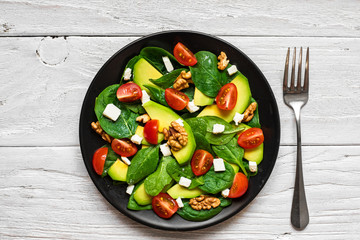 Fresh salad with avocado, spinach, tomatoes cherry, feta cheese and walnuts in a plate with fork on white wooden table