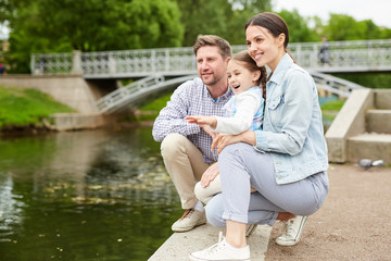 Family of three sitting by waterside in park and looking at birds while enjoying their chill