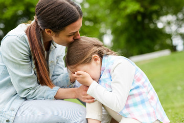 Young woman kissing her crying daughter on head, embracing and comforting her during chill in park