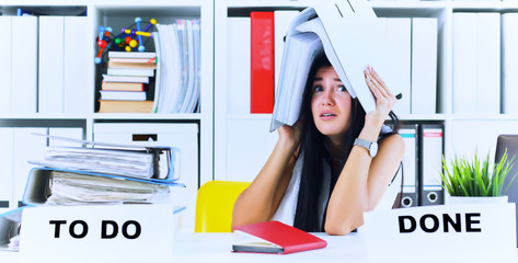 Funny overworked girl with a folder on her head sitting in the workplace cluttered with folders. Reaction of a subordinate when the boss orders her to do too much.