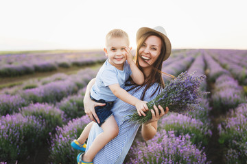 Young woman in blue dress hat walk on purple lavender flower meadow field background, rest, have fun, play with little cute child baby boy. Mother small kid son. Family day, parents, children concept.