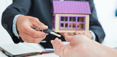 Real estate agent in suit holds house model in one hand and in the second hand holds out the keys of the house to woman.