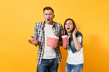 Young shocked scared couple woman man watching movie film on date holding bucket of popcorn and plastic cup of soda or cola spreading hands isolated on yellow background. Emotions in cinema concept.