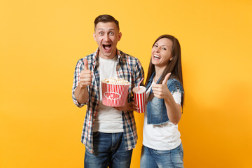 Young smiling laughing couple woman man watching movie film on date holding bucket of popcorn plastic cup of soda or cola showing thumbs up isolated on yellow background. Emotions in cinema concept.