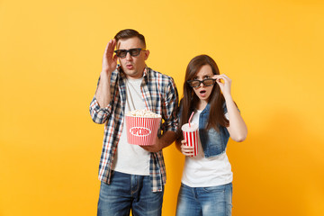 Young shocked couple woman man in 3d glasses watching movie film on date holding bucket of popcorn plastic cup of soda or cola keeping hand near face isolated on yellow background. Emotions in cinema.